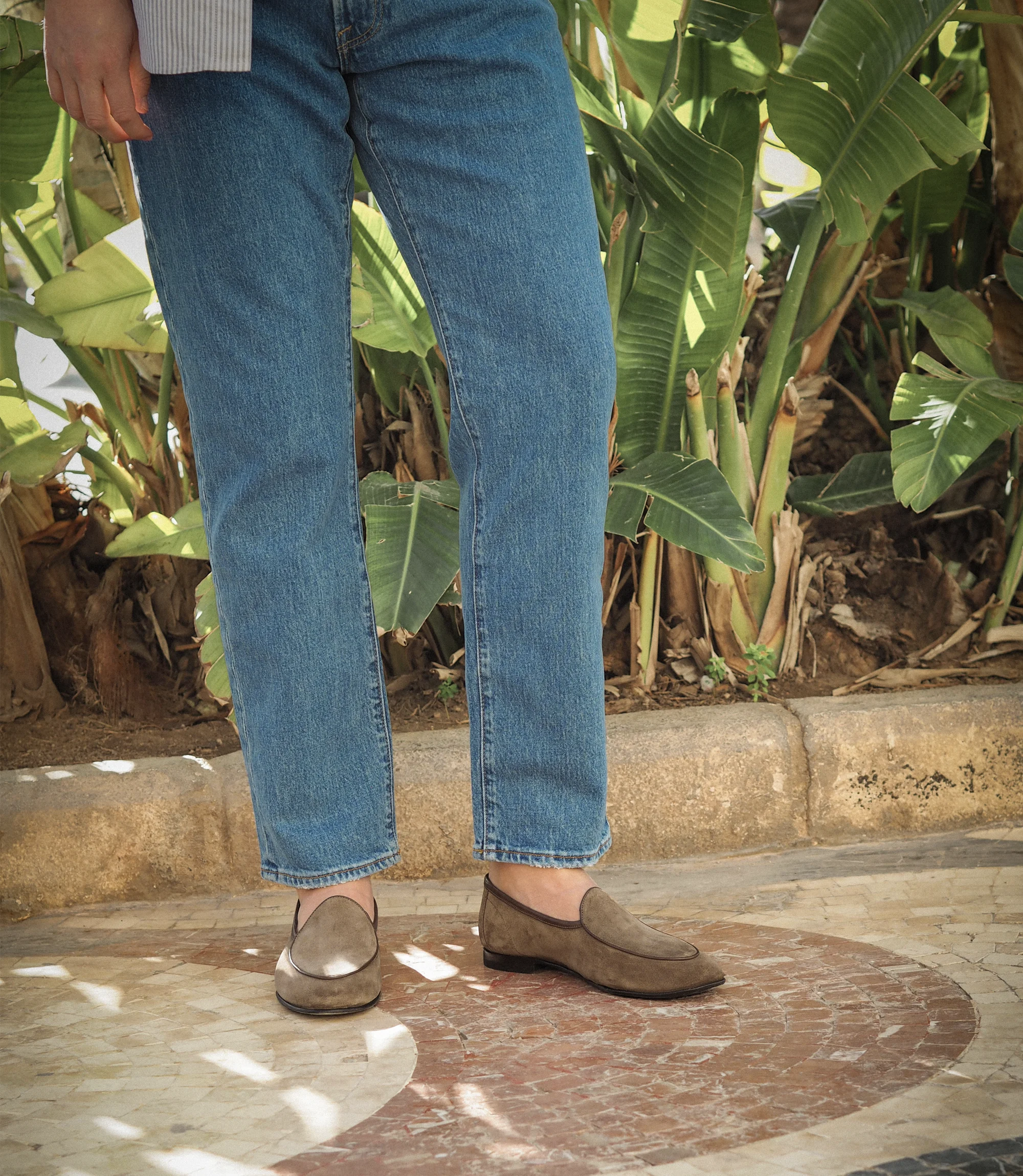 A man standing in front of a plant outside in navy blue jeans wearing his Loake Belgrave Loafers in Flint suede.