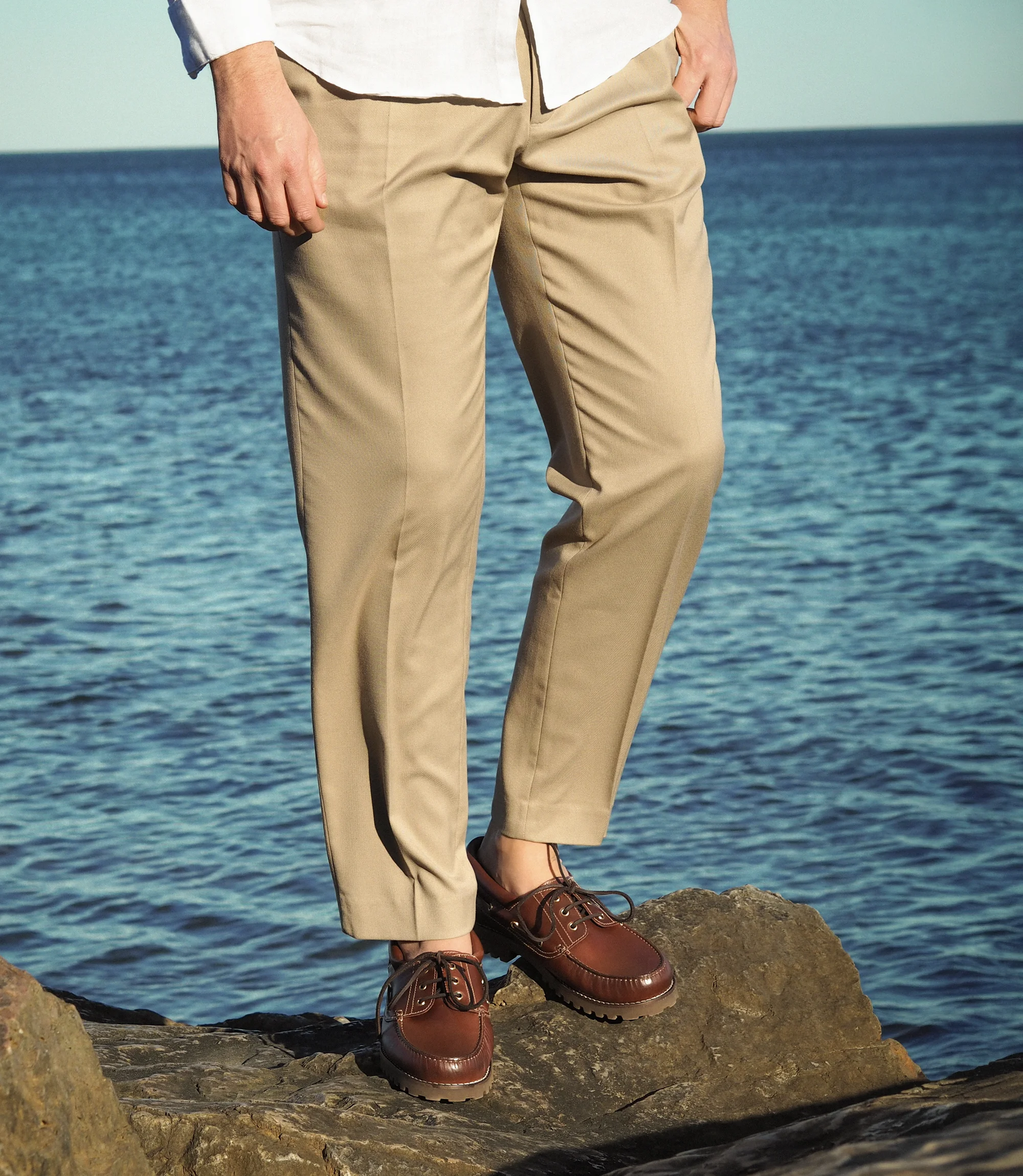 A man standing on rocks with the sea in background wearing smart beige trousers wearing his Loake 523 brown waxy Boat shoes.