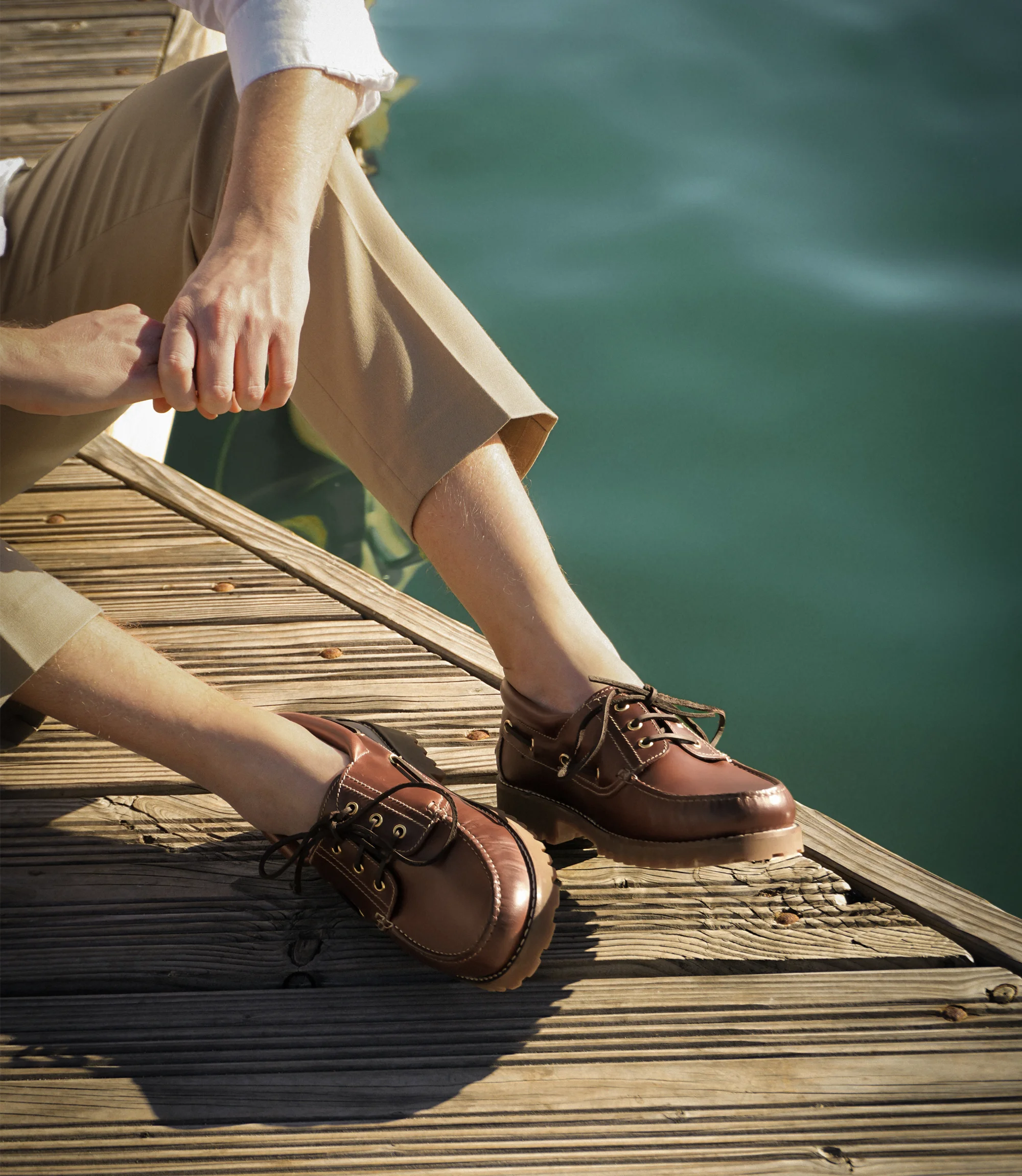 A man sitting down by the sea wearing smart beige trousers and a white shirt wearing his Loake 523 brown waxy Boat shoes.