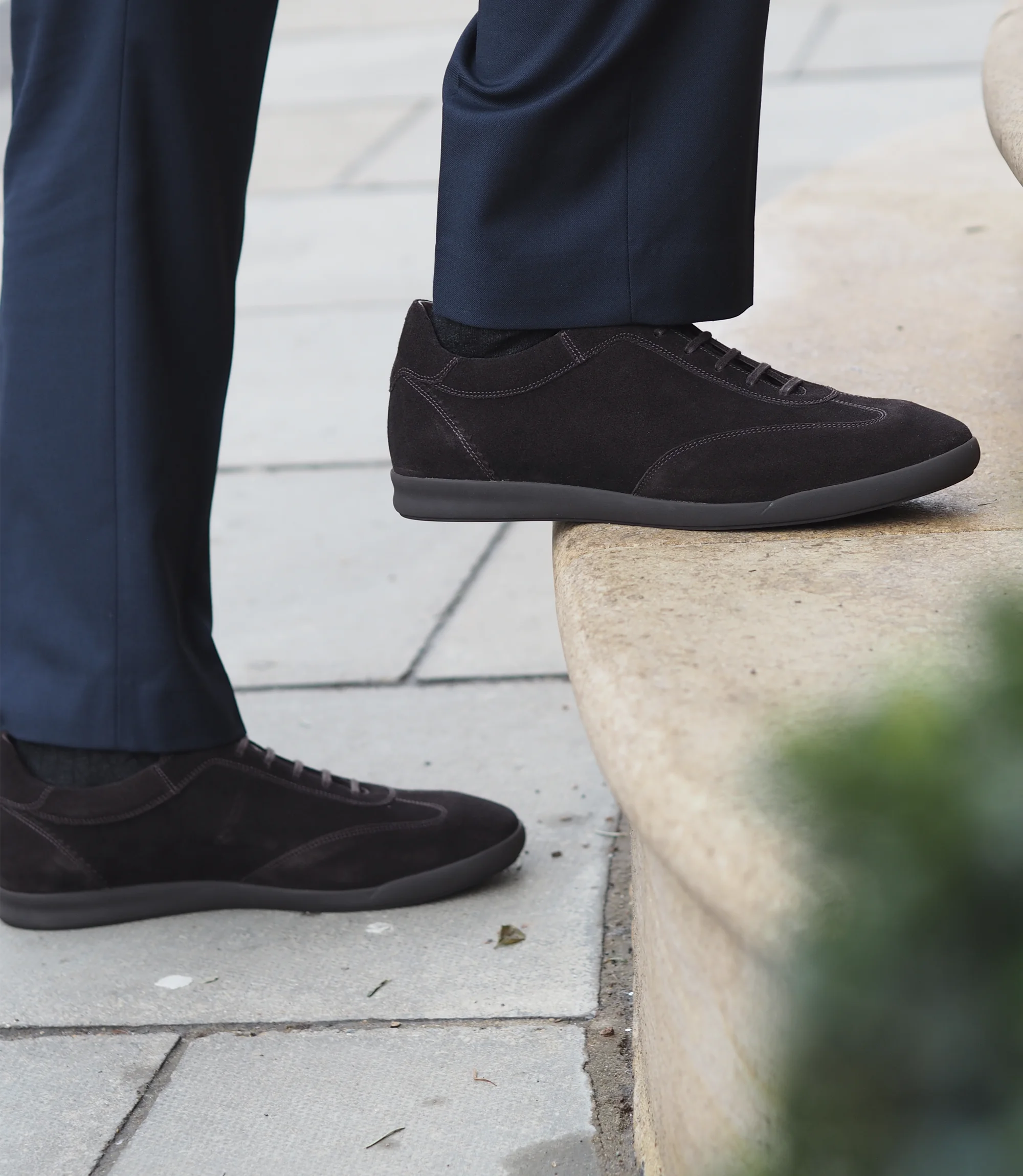 A man in a navy blue suit walking up stairs wearing his Loake Mayfair Smart Trainers in Dark Brown Suede.