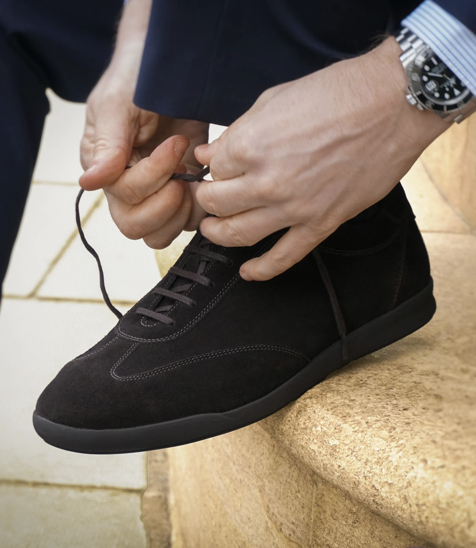 A man in a navy blue suit sitting on a step wearing his Loake Mayfair Smart Trainers in Dark Brown Suede.