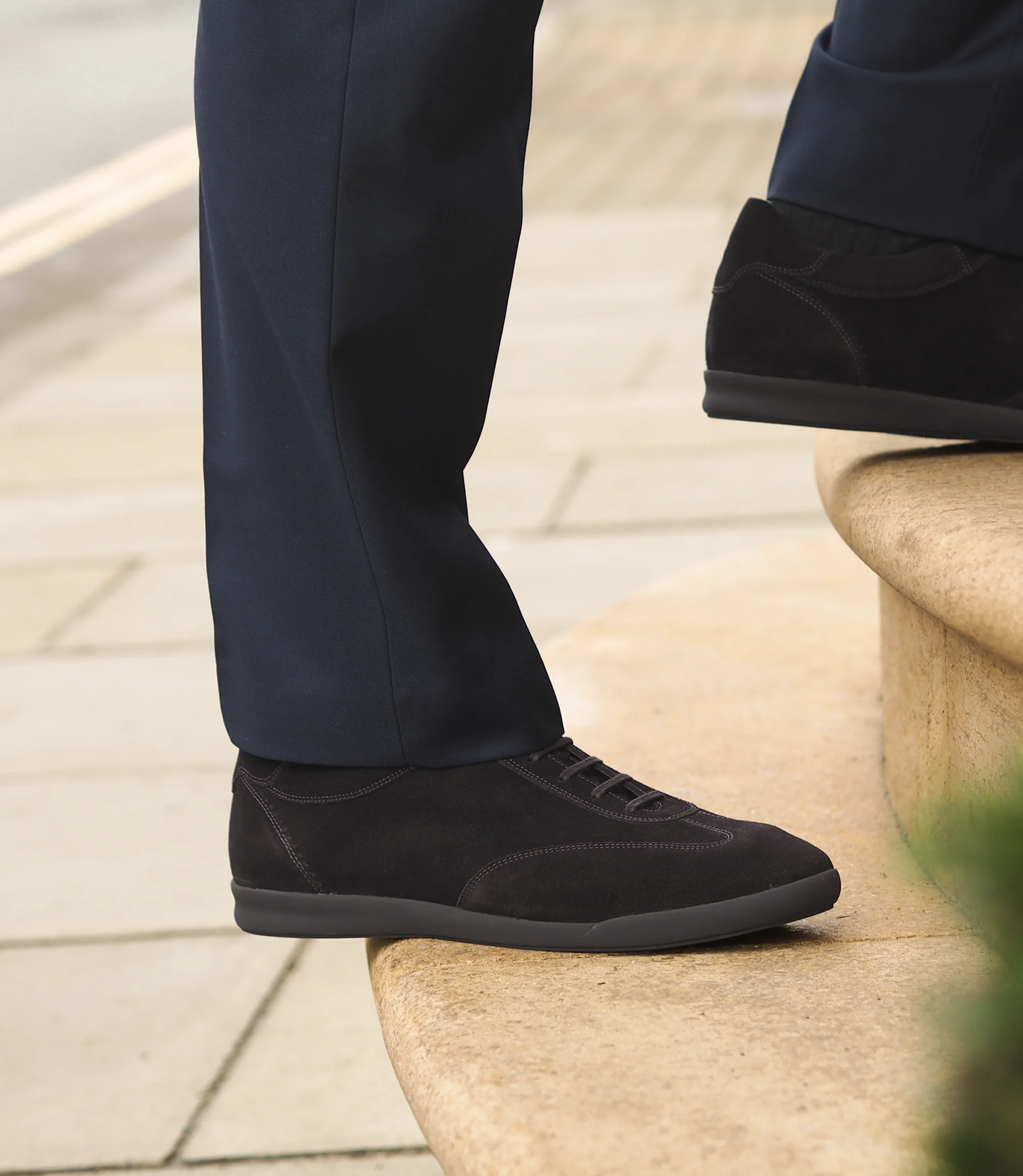 A man in a navy blue suit walking up stairs wearing his Loake Mayfair Smart Trainers in Dark Brown Suede.