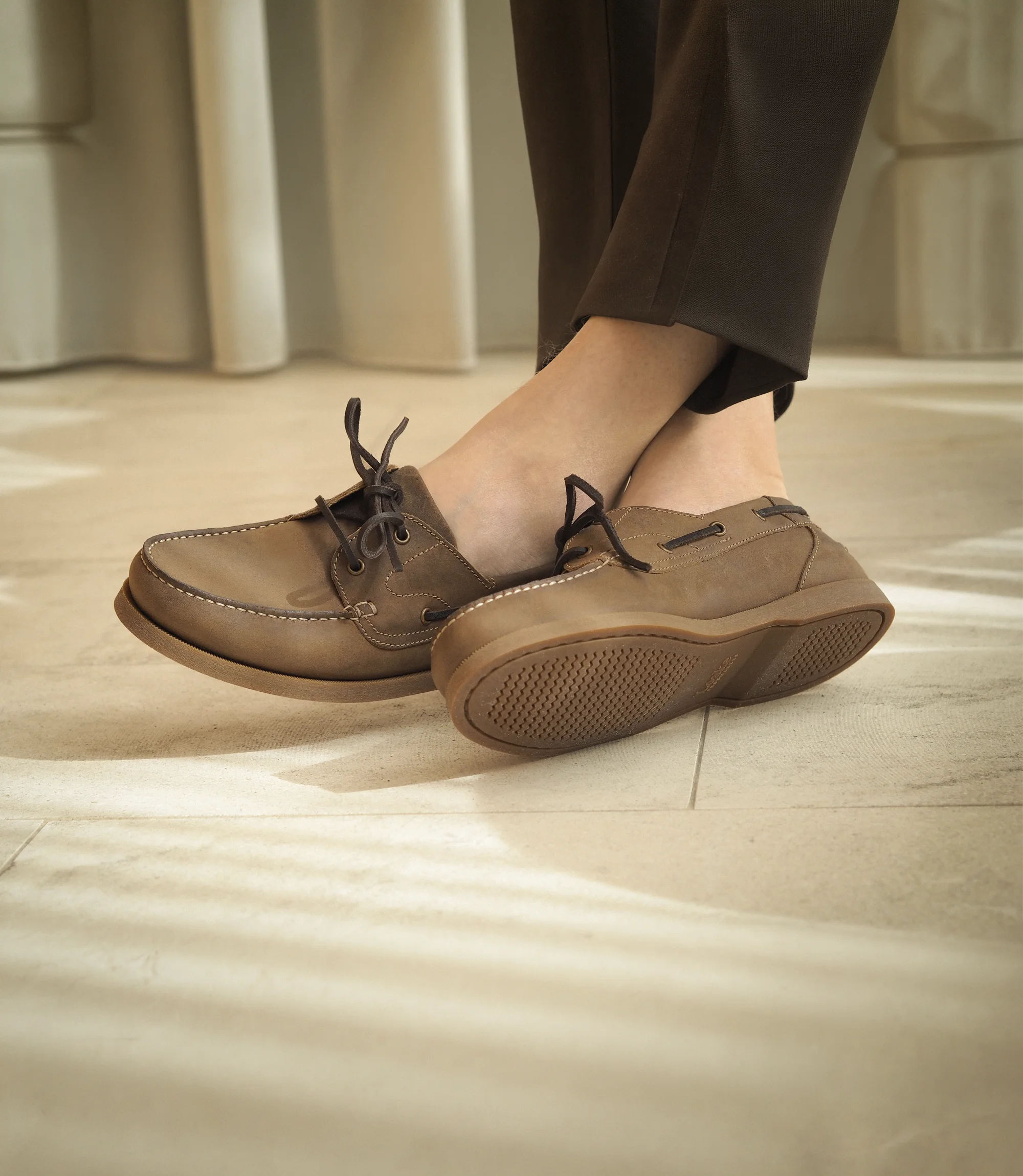 A man sitting on a wooden chair inside a building wearing smart brown trousers with crossed legs wearing his Loake Padstow Boat shoes in Brown Oiled Nubuck.