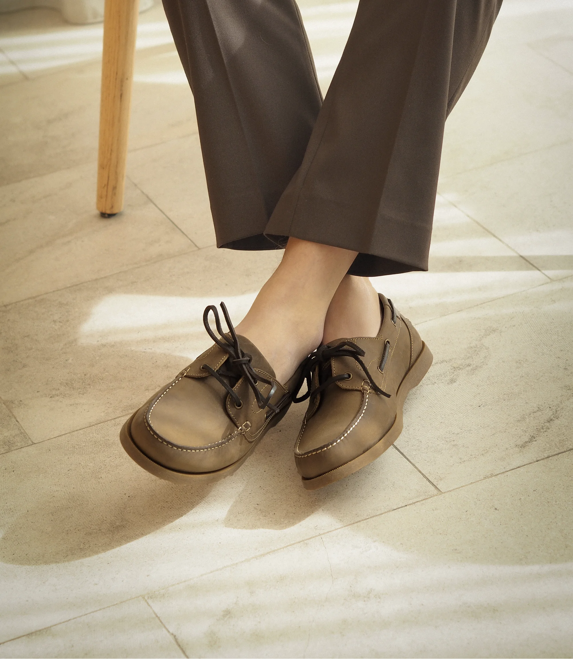A man sitting on a wooden chair inside a building wearing smart brown trousers with crossed legs wearing his Loake Padstow Boat shoes in Brown Oiled Nubuck.