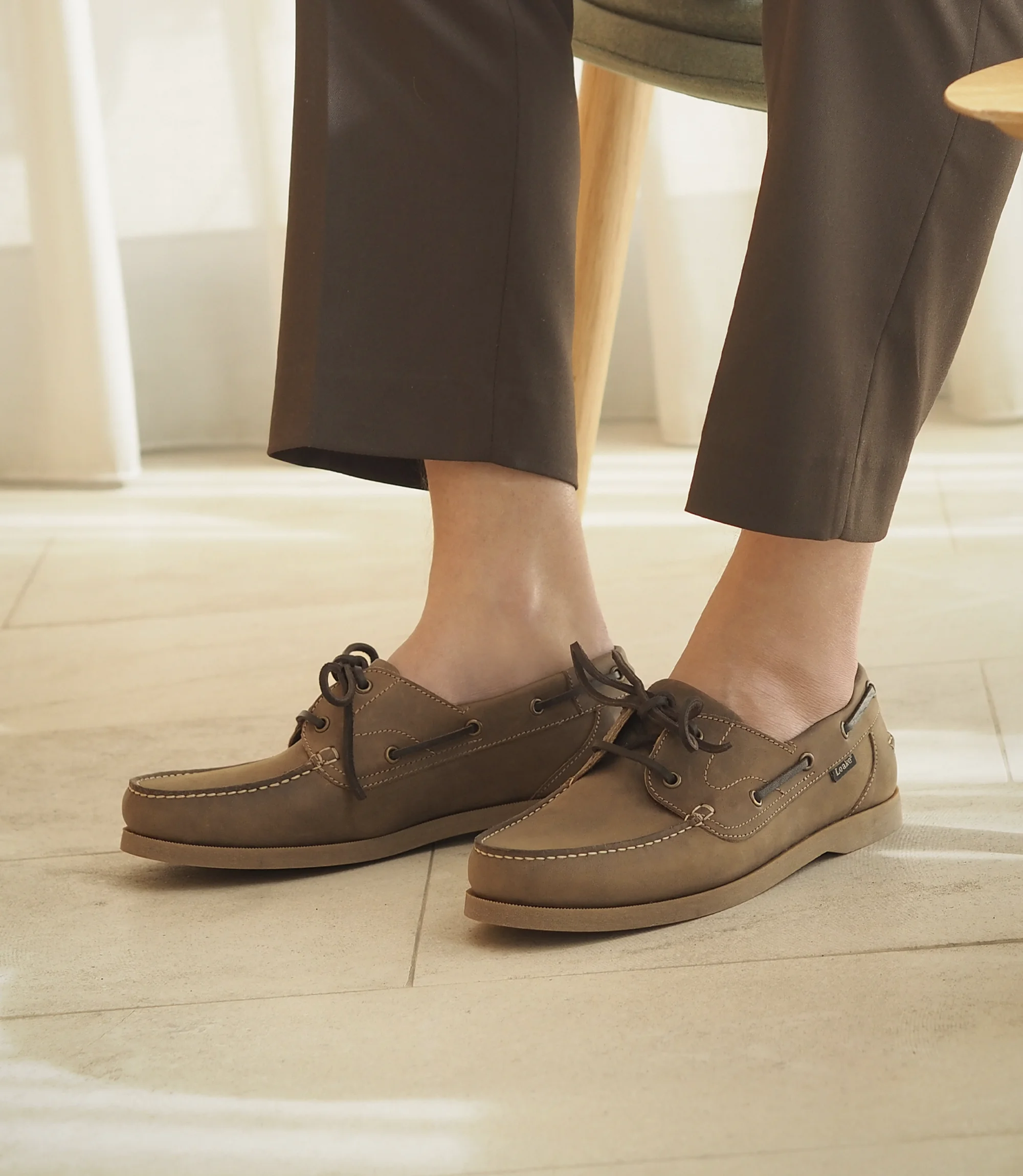 A man sitting on a wooden chair inside a building wearing smart brown trousers wearing his Loake Padstow Boat shoes in Brown Oiled Nubuck.