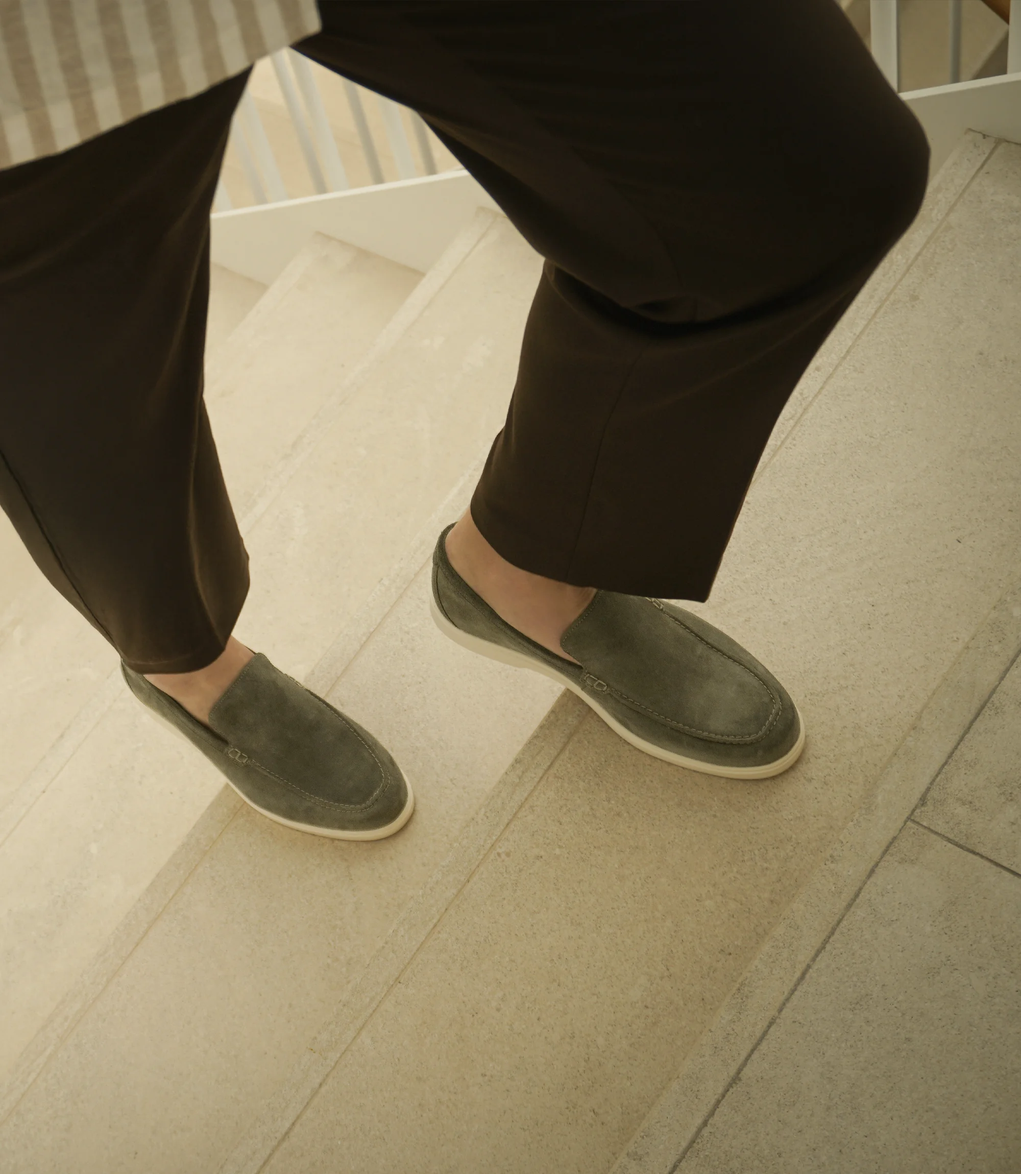 A man walking up stairs on a hotel building wearing brown smart trousers wearing his Loake Tuscany Loafers in Olive suede.