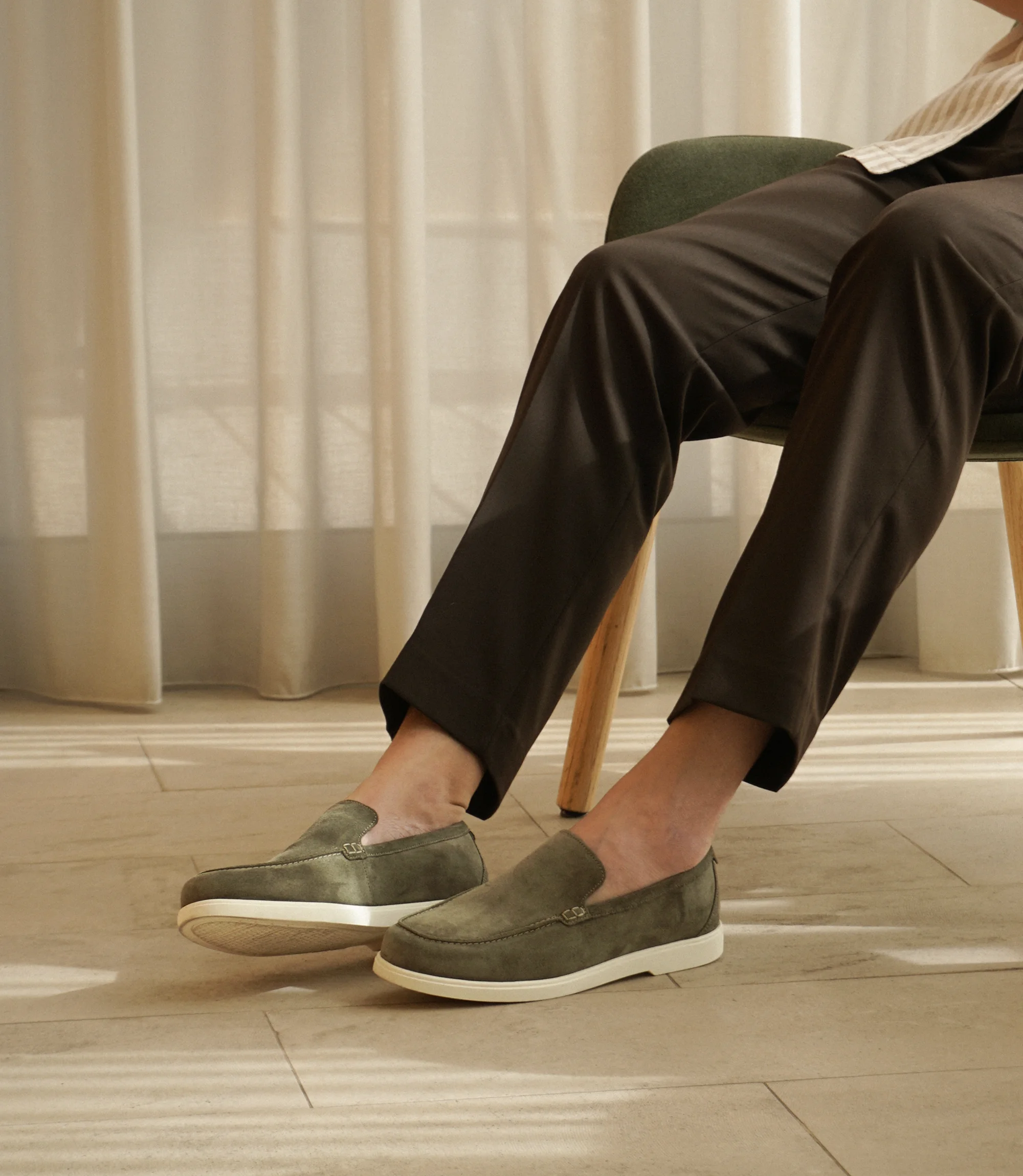A man sitting down on a chair wearing brown smart trousers wearing his Loake Tuscany Loafers in Olive Suede.